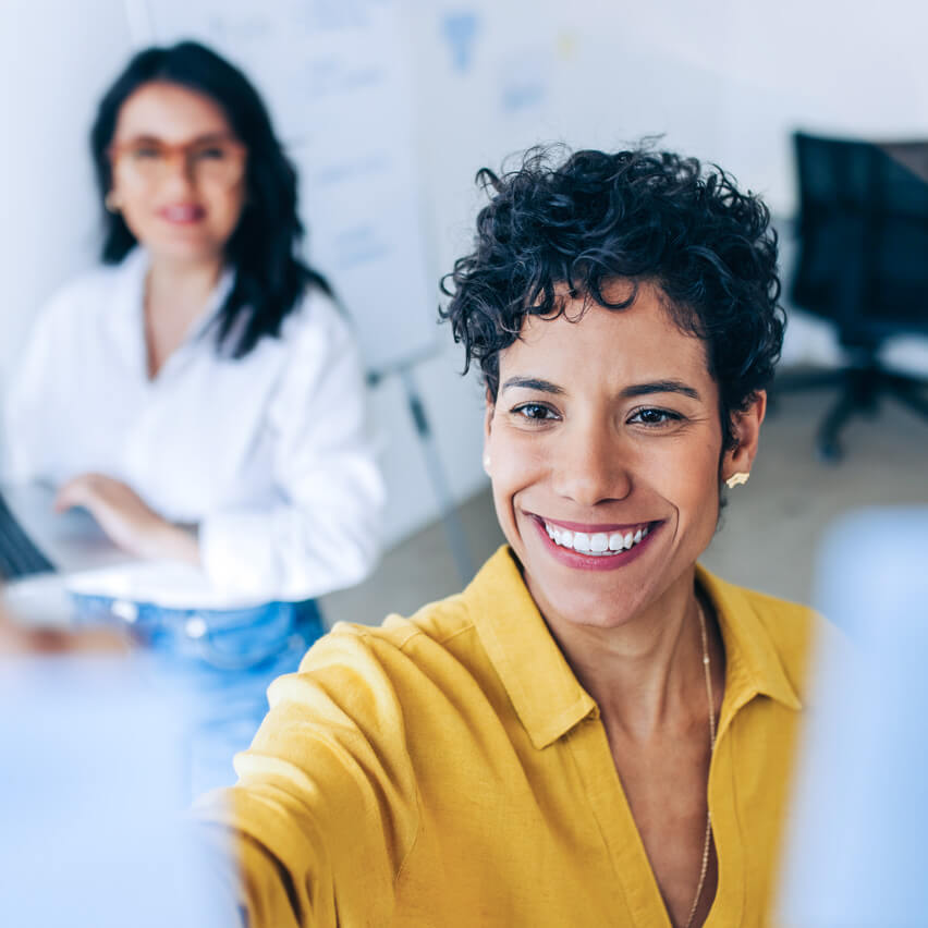 Frau in gelber Bluse lächelt in einem Workshop, im Hintergrund eine Kollegin.