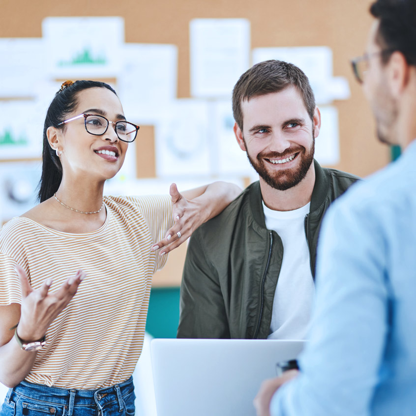 Drei junge Berufstätige im Gespräch vor einem Laptop, in lebendiger Teamatmosphäre mit Präsentationsmaterial im Hintergrund.