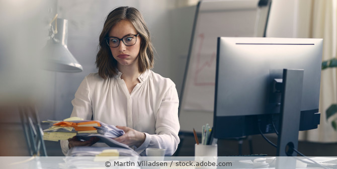 Angestellte sitzt am Schreibtisch und schaut verzweifelt auf einen Stapel Akten.